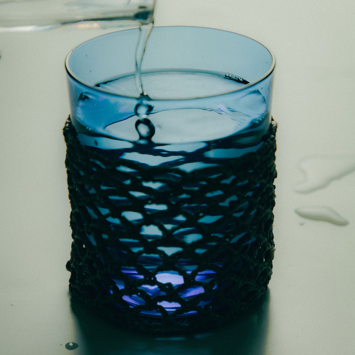 Blue glass of water with a woven raffia sleeve being filled on a white background