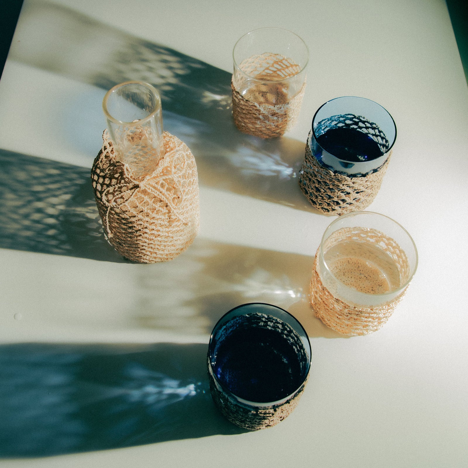 Four glasses and a carafe wrapped in raffia on a white surface